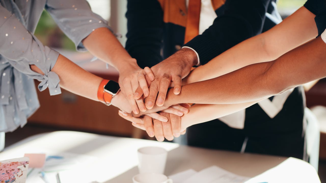 Home A close-up shot of diverse team members stacking hands over a white table, symbolizing unity and collaboration.